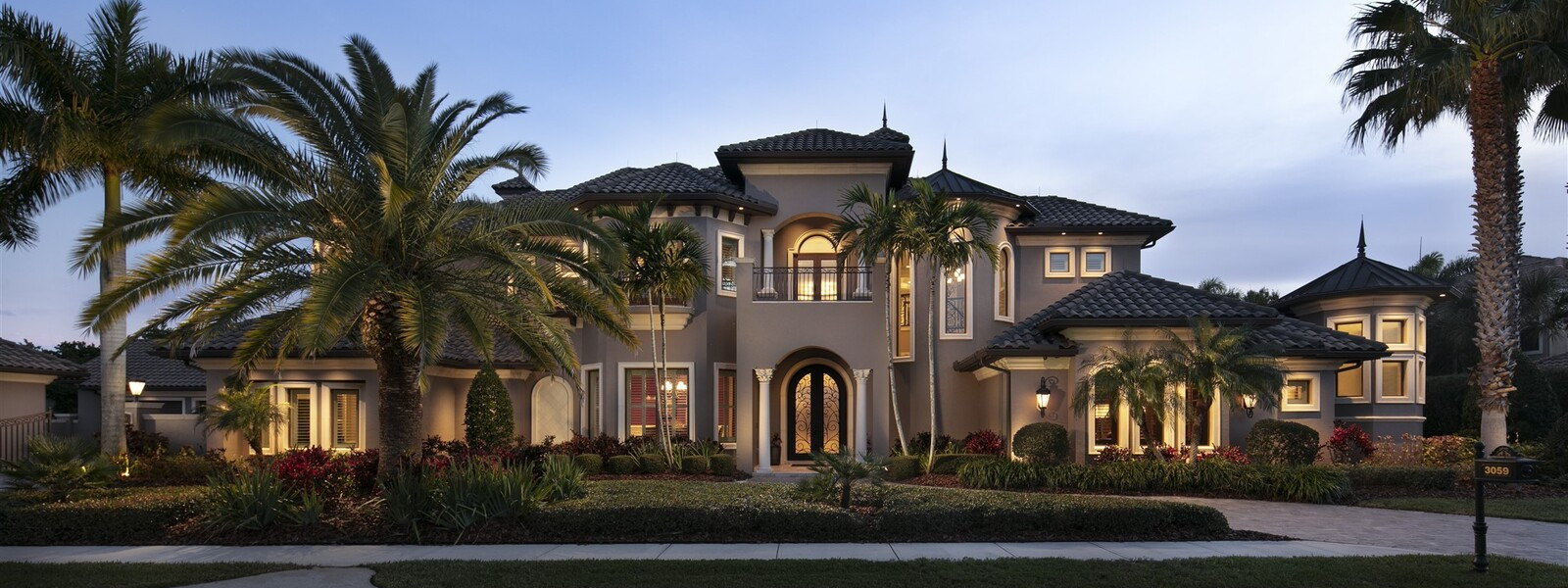 Front elevation of massive estate taken at twilight.  Palm trees in forground, light eminating from all windows and arched iron scroll double doors.  Home two story light brown with cream trim and columns.  Typical Brevard County, FL architecture